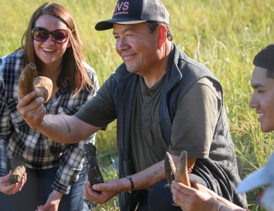 A group of three people outdoors, smiling and holding various objects, surrounded by tall grass.
